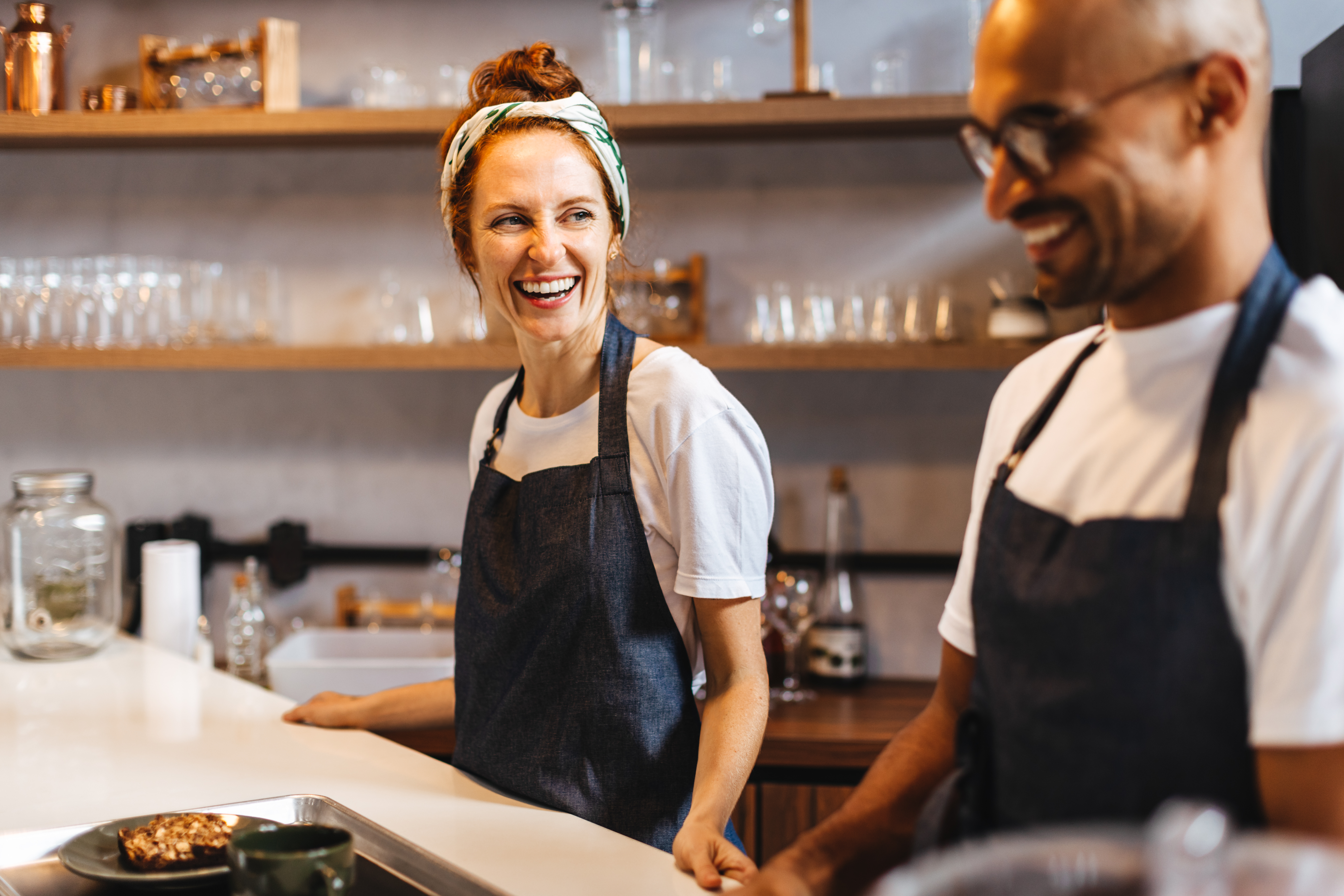 Two happy baristas stand side by side behind the coffee bar, preparing to serve customers. Waitstaff members preparing to brew up the perfect coffee and deliver exceptional service in a coffee shop.